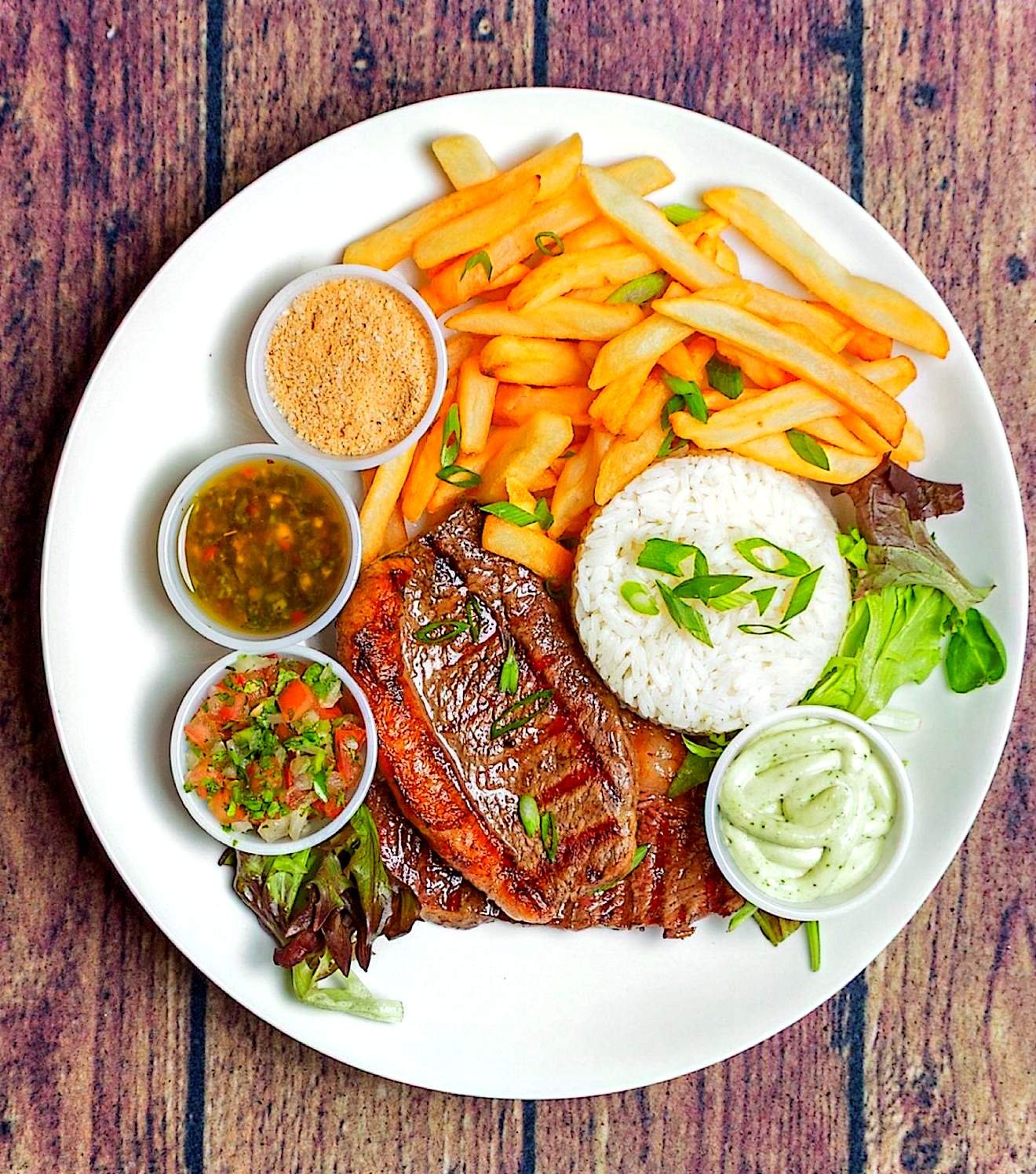 Grilled steak served with rice, fries, farofa, vinaigrette, and herb sauce